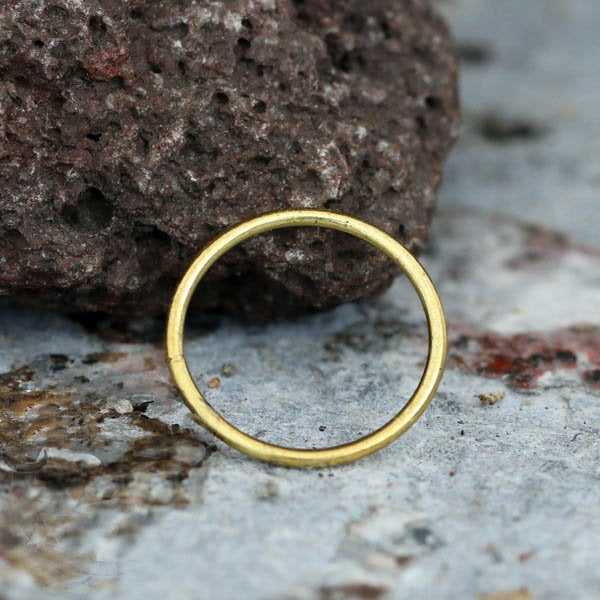 The INDUS Seamless Hoop Ring in Gold by Kaya Movement, a simple and slim 20-gauge piece, rests on a textured, rocky surface. The ring's smooth and consistent circular shape contrasts with the roughness of the background. The rocky surface displays a variety of earthy tones and textures.